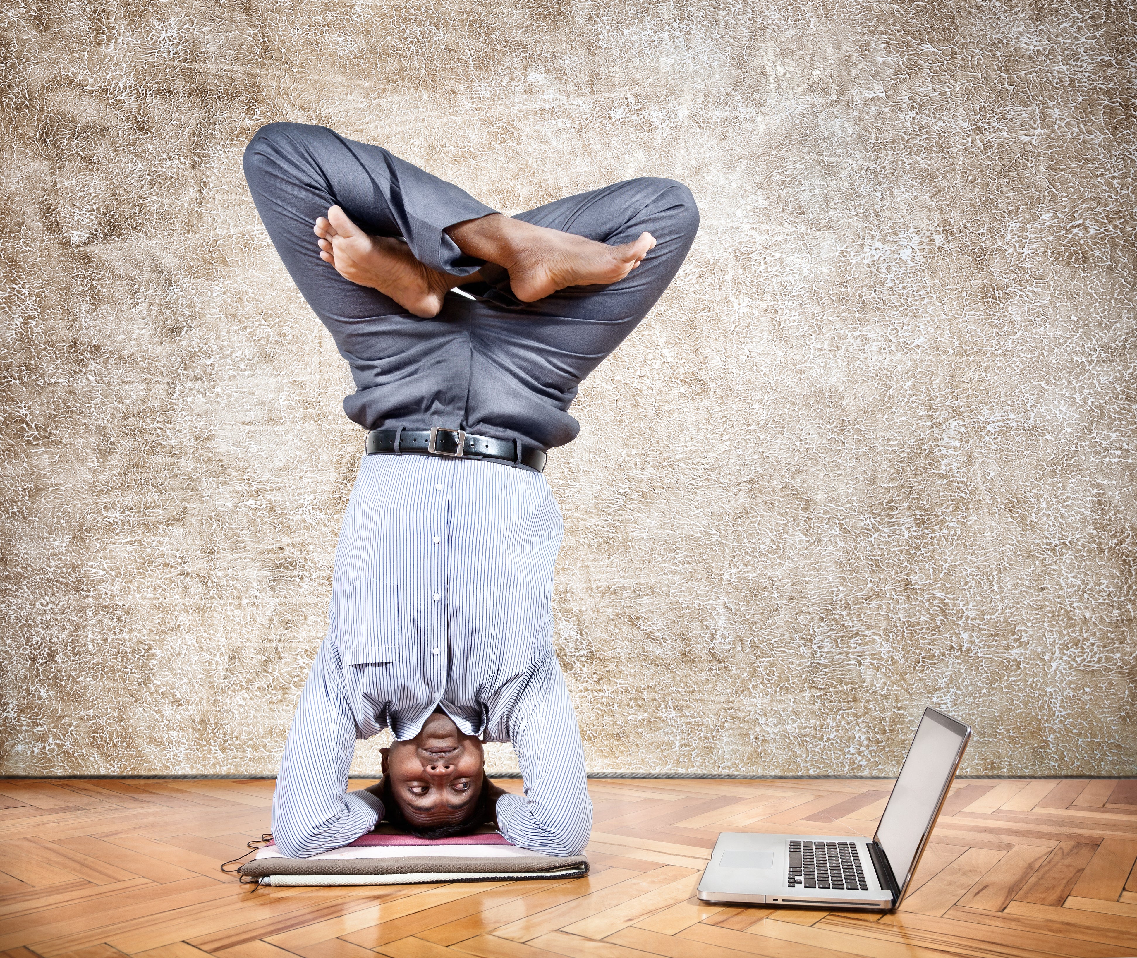 Man doing yoga pose head stand legs crossed, laptop next to him