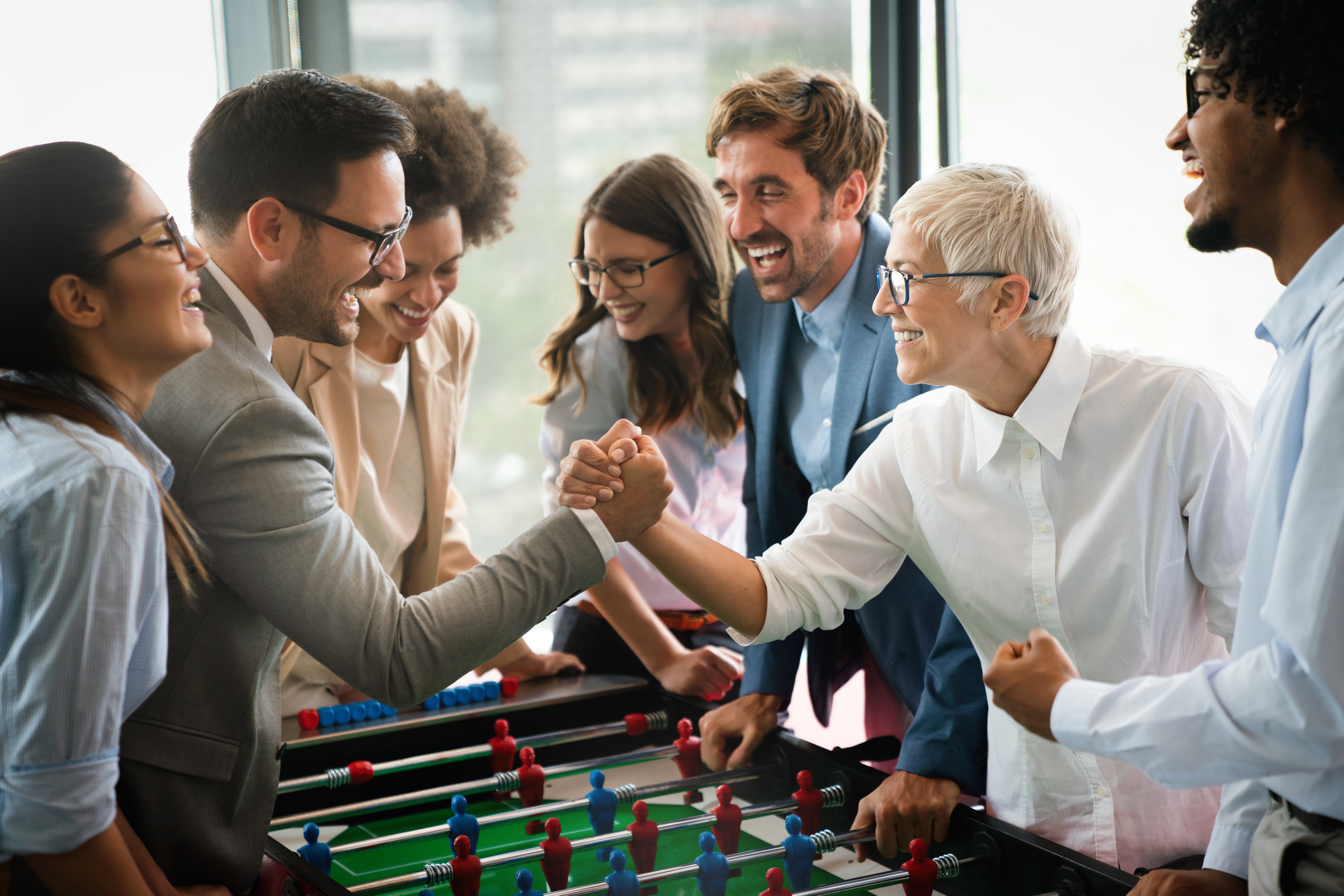 photo of group playing foosball, dressed professionally for office, one, appears to be woman, white, gray hair, dap (dignity and pride) handshake with man, brown, looking younger, surrounded by mixed group
