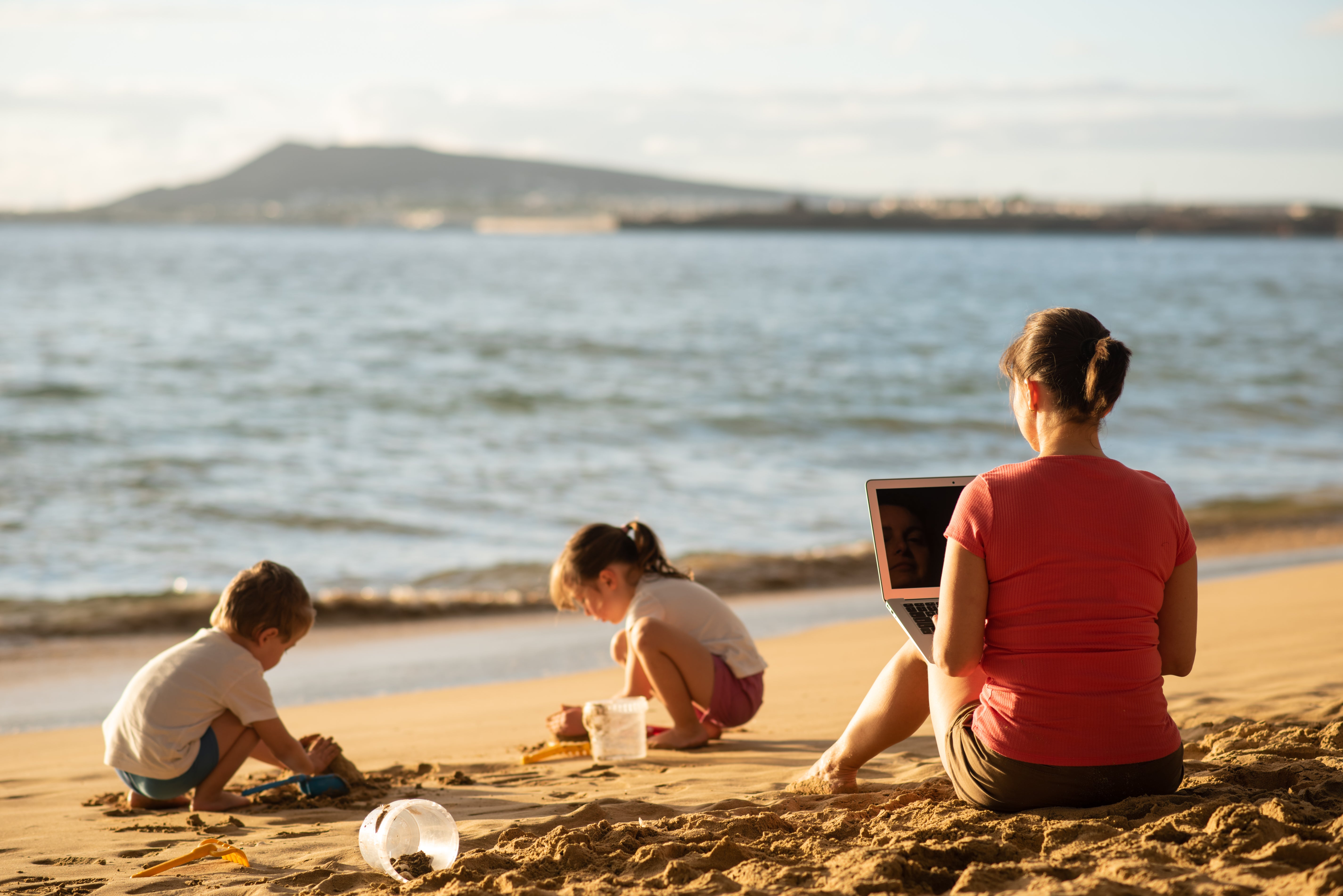 A woman working remotely at the beach with two children. 