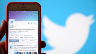 A phone with the twitter profile on screen with the Twitter logo in the background