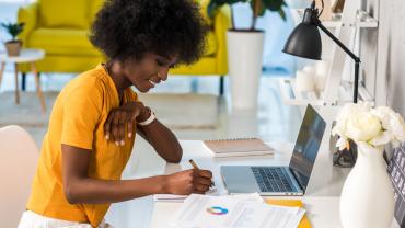 Happy African American business woman smiling and wearing a yellow shirt while sitting at a home office desk in front of a laptop next to white flowers and a yellow couch in the background.