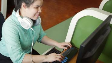 woman, brown or white, it appears, sitting at desk using computer, has headphones and a non-standard keyboard, may be for braille