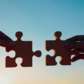 Silhouette of two hands putting two puzzle pieces together in front of a blue and yellow sky at sunset.