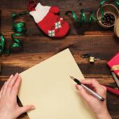 A hand poses with a pen to write on a blank piece of paper. On a table surrounded by Christmas wrapping materials.