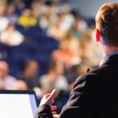 man at a podium, with back facing the viewer, speaks to an audience in the background