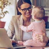 a baby is seated on a desk, held by her smiling mother while simultaneously works on her computer