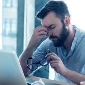 man seated at his computer with his glasses off holding the bridge of his nose