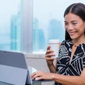 seated young female professional smiling while typing at her computer with her left hand and holding her coffee in the right hand