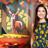 profile photo of smiling Sarah Feingold standing in front of an oil painting of family of deer