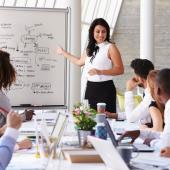 female leader standing next to dry erase board talking to her seven person team seated at a table