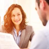 a female professional is facing the viewer with a male's back to the viewer holding papers during an interview