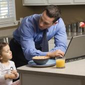 a father in his shirt and tie at the kitchen island leans over his computer looking at his daughter eating breakfast