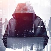 hooded figure, face not showing, hulking over laptop, figures and buildings in background, as though hooded one is hacking into their network
