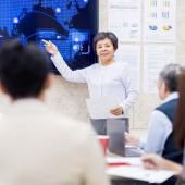 photo of, appears to be Asian woman, gray in hair, addressing group sitting, mixed ages, gender, culture, looking smartly business casually dressed, pointing at screen, smiling