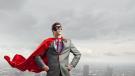 man (appears to be, white), in suit, wearing red cape like Superman type, eye mask, cape floating like he might have flown or will fly, skyline in background like he's above it